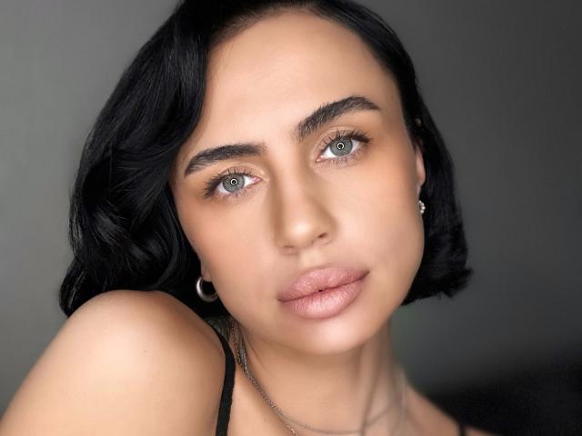 LeaNoir has short black hair, light brown skin, green eyes, and full pink lips. She's wearing small hoop earrings and a black strap, looking directly at the camera.