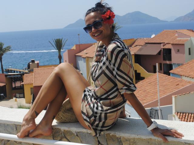 LeaNoir with dark skin, wearing a plaid dress, black sunglasses, and red hibiscus in hair, sitting on a stone ledge, smiling, showing her bare ass and toes.