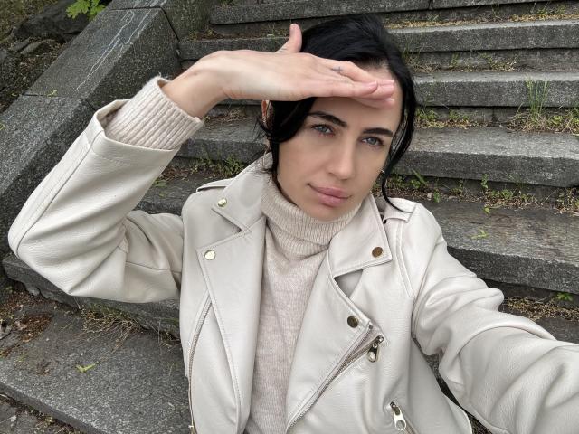 LeaNoir with dark hair, pale skin, wearing a beige leather jacket and turtleneck, covering her eyes with her right hand, standing on stone steps with grass.