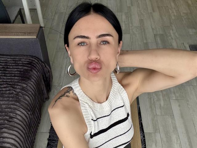 LeaNoir with black hair, pouty lips, wearing a striped sleeveless top, silver hoop earrings, tattoo on right arm, and arm raised behind head, on wooden floor.
