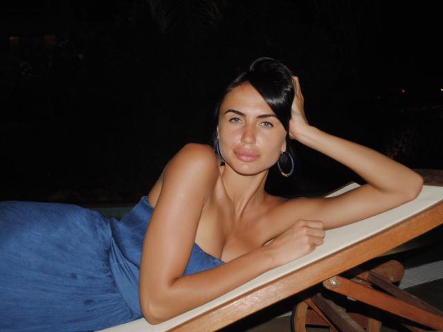 LeaNoir is lying on a wooden lounge chair, wearing a blue strapless dress, with her hand on her head, showing her tits and a relaxed, sexy expression.