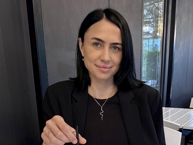 LeaNoir with black hair, blue eyes, wearing a black blazer, and a wavy necklace, smiling slightly, holding a pen, in a modern office setting.