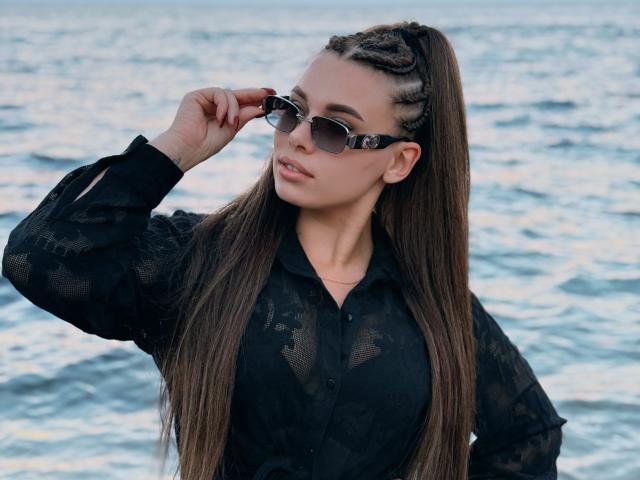 Niferttiti with long brown braided ponytail, wearing black lace shirt, adjusting mirrored sunglasses, standing by blue ocean, showcasing her tits and serious expression.