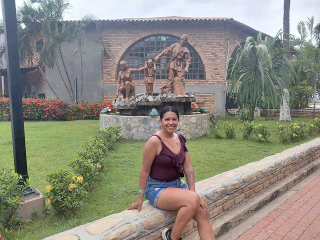 PauletteLoveX sits on a stone wall in the foreground. She has a medium build, dark hair in a ponytail, and wears a purple sleeveless top with denim shorts. She smiles at the camera. Behind her, a bronze statue of four children stands on a stone pedestal, surrounded by greenery and red flowers. The building in the background has a brick facade with a large arched window. Palm trees and a tall street lamp are to the left. The ground is a mix of grass and brick.