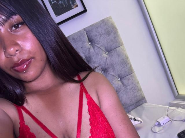 NahomyNoir, com a pele marrom brilhante, cabelo preto liso e um sutiã de renda vermelho, mostrando seus pequenos seios e lábios rosados e levemente entreabertos, sentada em uma cama cinza com estofamento..