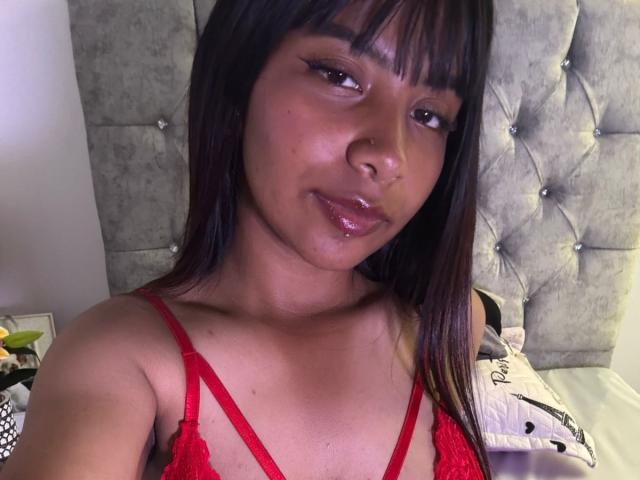 NahomyNoir has shiny brown skin, dark straight hair with bangs, red lace bra, and glossy lips, looking at the camera with a slight smile, showing her tits.
