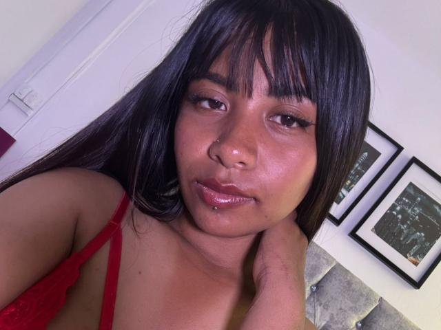 NahomyNoir with dark straight hair, bangs, nose ring, glossy pink lips, red lace bra, hand on neck, brown eyes, slight smile, smooth skin, soft lighting, casual selfie.