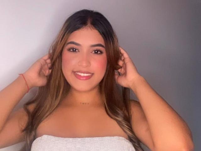 SusanaKloe has long brown hair, a cute smile, and is topless with her hands in her hair, showing her small tits and a red bracelet on her left wrist.