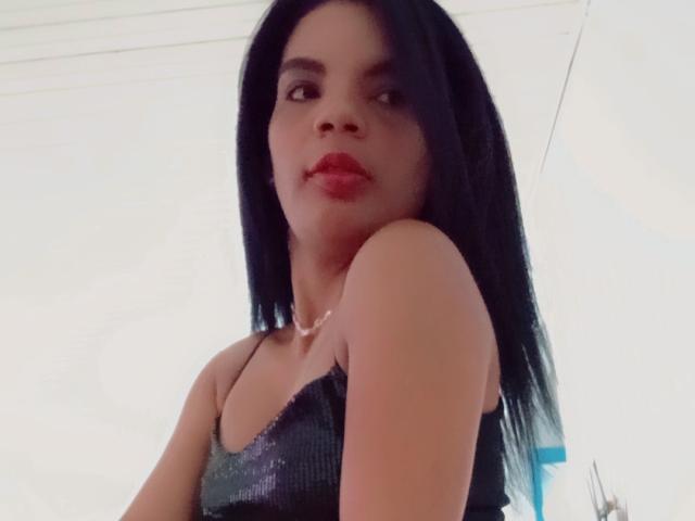 RebecaEla with black hair, red lipstick, wearing a sparkly black dress, looking down, showing one bare shoulder, subtle necklace, soft light, slightly blurred, casual selfie.