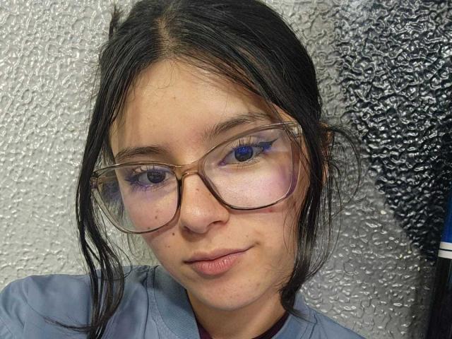 EmiSchwarz has light brown skin, black hair in a messy ponytail, and wears clear glasses with purple eyeliner. She has small, unobtrusive tits and a slight, confident smile.