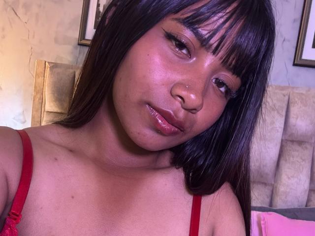 NahomyNoir has shiny brown skin, dark straight hair with bangs, and is wearing a red bra. Her nose is pierced, and she's giving a slight pout with glossy lips.