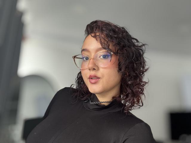 SophiaBrowm with curly dark brown hair, wearing glasses and a black sheer top, showing her boobs and pink lipstick, looking at the camera with a slight smile.
