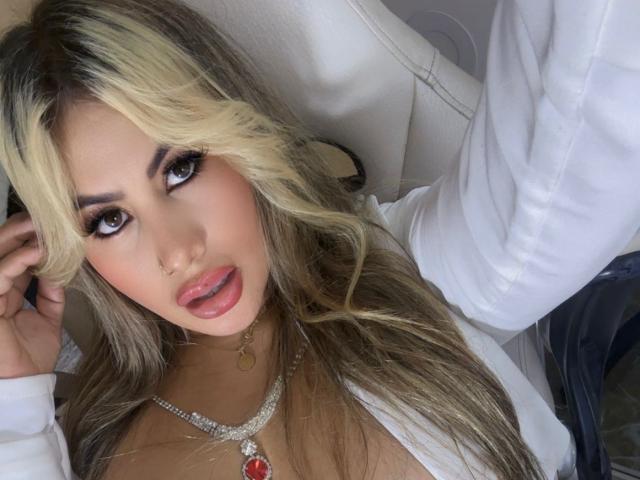 ChanellBrad with blonde hair, heavy makeup, and pink lips, wearing a white shirt, necklace, and red pendant, looking up with slightly open mouth, showing her tits.