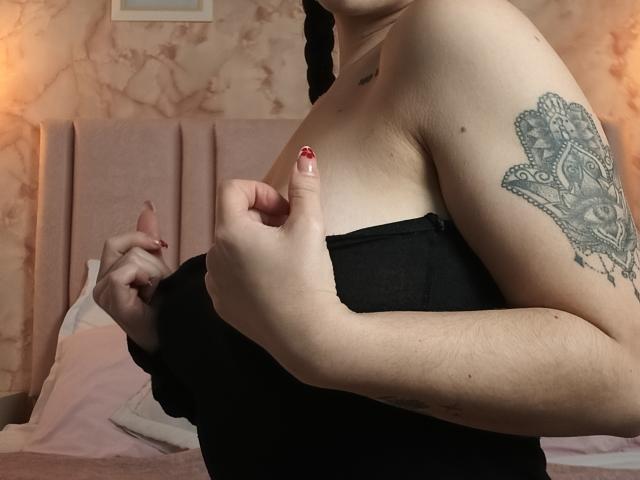 AnntonellaWatson with pale skin, black strapless top, visible tattoo of an intricate eye on her upper arm, and red manicured nails, touching her small tits with her right hand.