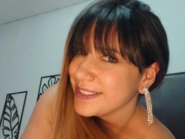 RoxanneBlair has dark brown hair with bangs, wearing a diamond chandelier earring, smiling with pink lipstick, and showing her bare shoulders. Her brown eyes are slightly upturned.