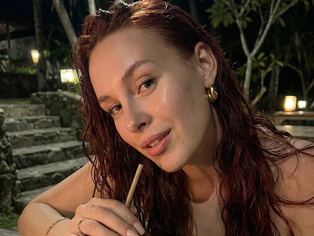 CleoErgaX with wet red hair, smiling, holding a straw, wearing gold earrings, has fair skin, and is topless in a dimly lit, outdoor setting.
