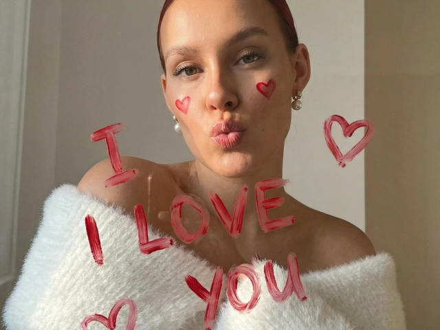 CleoErgaX with red heart marks on cheeks, pouty lips, red hair, white fluffy off-shoulder top, pearl earrings, "I LOVE YOU" in red paint. Boobs partially covered by top.