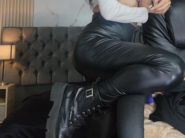 NenaBlack is seated on a black leather chair, her right leg crossed over the left. She wears black leather pants and black combat boots with metal buckles and laces. Her white rib-knit shirt is visible, but her face is out of the frame. The leather chair has a tufted, padded design. The bed in the background has a dark, textured blanket. The room has a modern, minimalist aesthetic with a lamp on a wooden nightstand. The lighting is soft, highlighting the textures of the leather and the rib-knit fabric.
