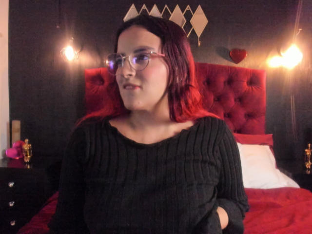 LaiaJonnes, a young woman with fair skin and red-dyed hair, wears glasses and a black ribbed sweater. She stands in front of a red tufted headboard, with a black wall in the background. Two hanging bulbs illuminate her. The headboard has geometric diamond decorations and a red heart. Her expression is neutral, and she has her hands in her pockets. The composition uses warm colors and contrasts the dark wall with the red headboard.