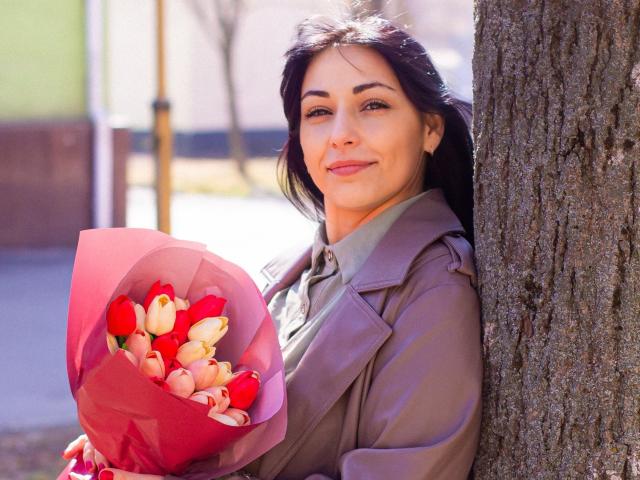 LiluGrants, dark-haired, smiling woman in a brown coat, holding a pink bouquet of red and white tulips, leaning against a tree, sunlit, with a relaxed, cute face.