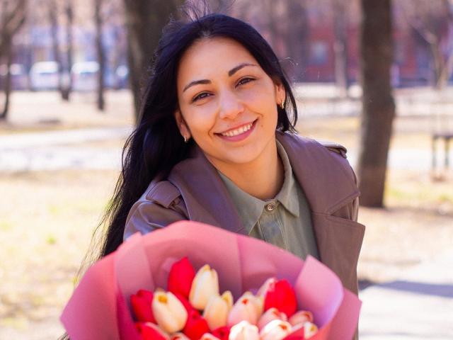 LiluGrants with long black hair, smiling, holding a pink bouquet of red and white tulips, wearing a brown jacket over a green shirt, in a sunny park.