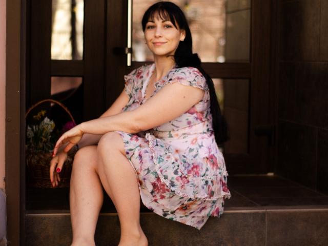 LiluGrants, sitting on a porch, wearing a floral dress, showing her tits in a semi-sitting pose, with long black hair and a basket of flowers behind her.