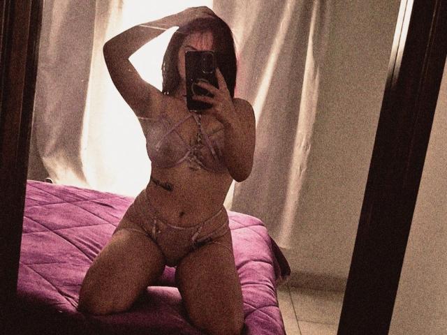 A mirror selfie of AshleyPlatta in a dimly lit room. She kneels on a purple quilted bed, wearing sheer, light pink lingerie with lace details. Her right hand touches her dark, shoulder-length hair, while her left hand holds a smartphone, partially covering her face. She has a small tattoo on her right side, near her waist. The room has beige curtains in the background, and the image has a slightly grainy texture. Her body is curvy, with a medium build and light skin.
