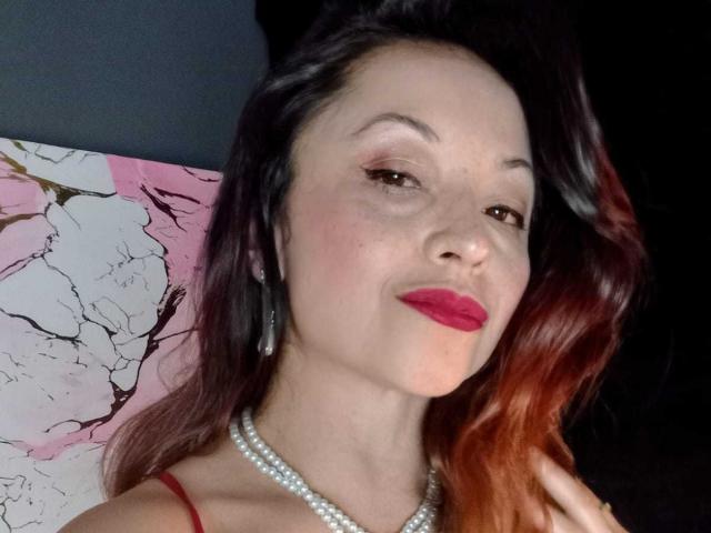 AngelinaBlanche has pale skin, red lipstick, and dark wavy hair with orange tips. She's wearing a pearl necklace and red strap, looking at the camera with a slight smile.