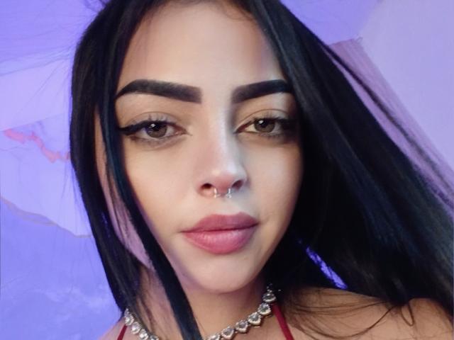 LilithAmoth has black hair, thick eyebrows, brown eyes, and a septum piercing. She's wearing a red top with a silver chain necklace. Her lips are pink and full.