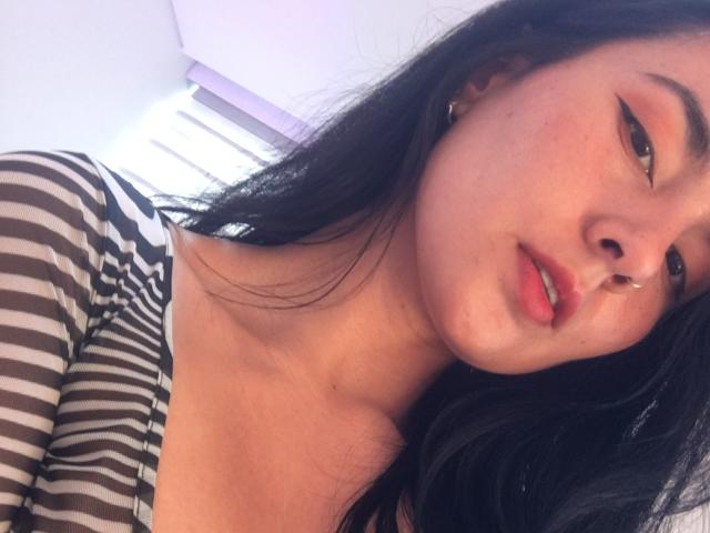 Close-up photo of Natii69, a young woman with long black hair, wearing a striped shirt. She has light brown skin, a small nose ring in her right nostril, and wears subtle makeup with pink lipstick. Her lips are slightly parted, and she looks to the left. Her striped shirt has black and white horizontal lines, and her collarbone is visible. The background is blurred and white. Her left ear has a small gold hoop earring. The image has a casual, slightly tilted composition.