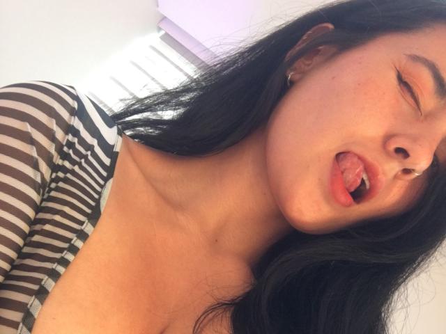 Close-up of Natii6 9 with long black hair, eyes closed, and tongue slightly out. She wears a striped black and white shirt, revealing a bit of cleavage. Her skin is light with some freckles, and she has small earrings. Her tongue is visibly protruding, and her expression is playful. The lighting is bright, and the focus is on her face and upper chest. The image has a casual, selfie-like quality.