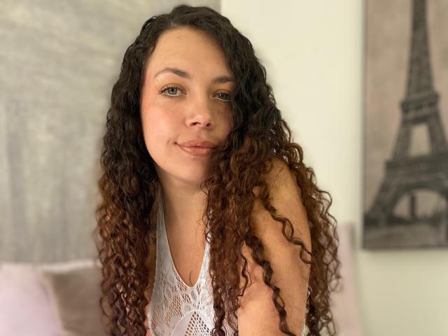 Close-up of RubbyStrongX with long, curly brown hair, wearing a white lace halter top. She has light brown skin, green eyes, and a slight smile. Her hair transitions from dark brown at the roots to lighter brown at the ends. The background includes a blurred, grayscale Eiffel Tower painting. The image has a soft, natural light, emphasizing her relaxed expression and casual pose. RubbyStrongX's subtle makeup and natural look are highlighted by the soft, warm color palette.