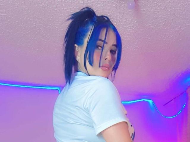 SofiaLeal with blue hair in a high ponytail, looking over her shoulder. She wears a white t-shirt, with a neutral expression. Her face has soft features and she has visible eyeliner. The background has pink and purple lighting, with blue neon lights on the ceiling. The texture of the ceiling is slightly rough. The image has a casual, modern style.