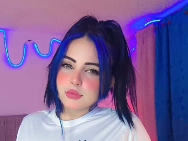 SofiaLeal with blue hair in pigtails, nose ring, and pink blush, wearing a white shirt, pouty lips, and blue neon lights in the background.