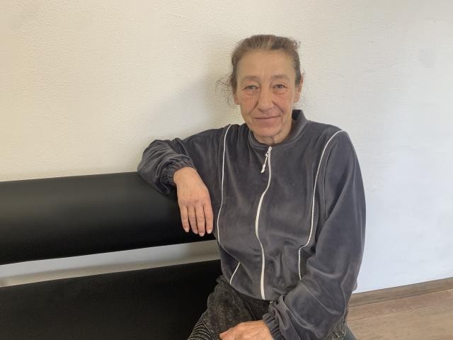 AngemlaMolda with gray hair, wearing a dark gray zip-up jacket, sitting on a black bench, smiling, right arm resting on bench, left hand on lap, wood floor.