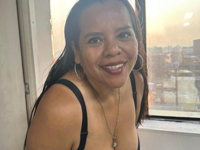 VeraCruz, a medium-skinned woman with long dark hair, stands near a window. She wears large silver hoop earrings, a black spaghetti strap top, and a gold necklace with a small pendant. She smiles widely, showing white teeth and dark purple lipstick. Sunlight from the window highlights her face and upper chest. Her expression is friendly and relaxed. The background includes a blurred cityscape through the window. The image focuses on VeraCruz's upper body and face.