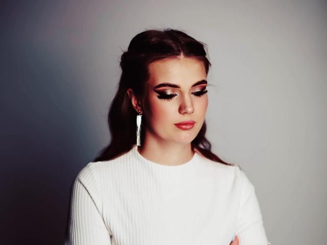 AdeleLily with long brown hair, white ribbed shirt, nose ring, and long earrings, looking down with dark eyeliner and pink lipstick, soft light on her face.