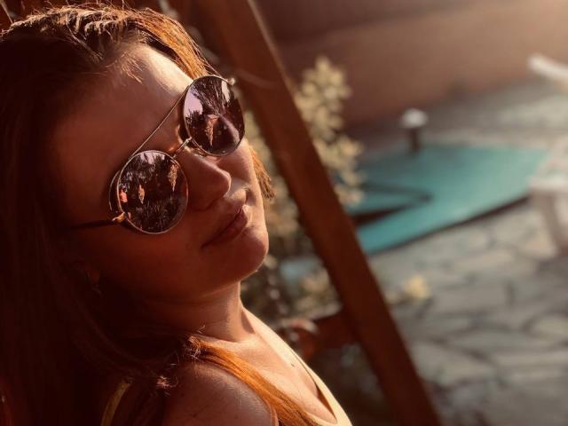 AdeleLily with reflective aviator sunglasses, orange hair, and a smile, sunlight highlighting her face and shoulders, wearing a yellow top, looking over her shoulder.