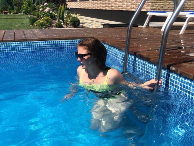 AdeleLily with tits in a green swimsuit, wearing sunglasses, climbing out of a blue-tiled pool with a metal ladder, her wet ass visible, brown hair, sunny day.