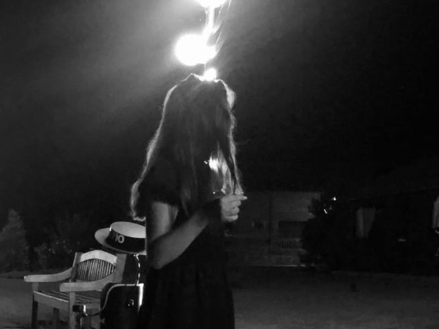 AdeleLily with long hair, holding a glass, wearing a dark dress, standing in front of a bright light, next to a chair and hat in a black-and-white photo.