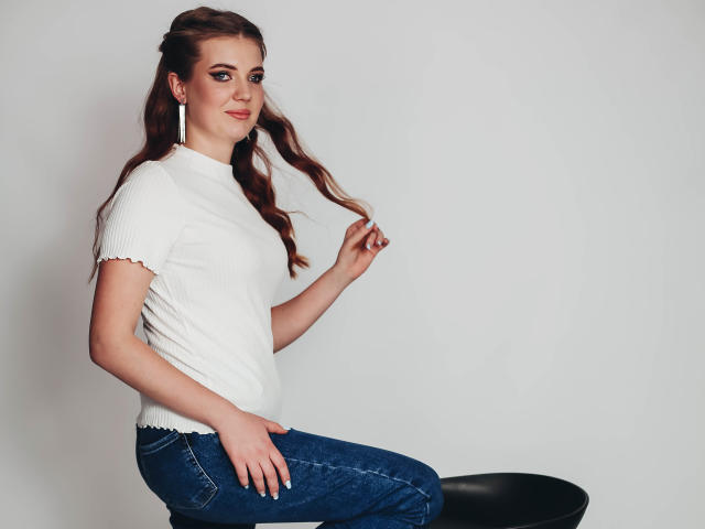 AdeleLily with wavy brown hair, white ribbed t-shirt, and blue jeans, sitting on a black stool, touching her hair, wearing long earrings, smiling slightly.