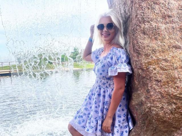 AdeleLily with long white hair, wearing a blue floral dress, stands against a rock, smiling, wearing sunglasses, water splashing in the background, touching her hair.