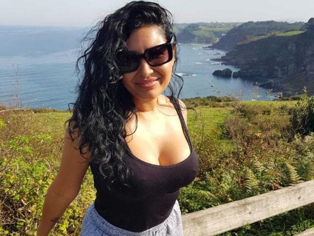 Naatyy with curly black hair, wearing black sunglasses, black tank top showing her big tits, and gray pants, standing by a wooden fence with a coastal view.