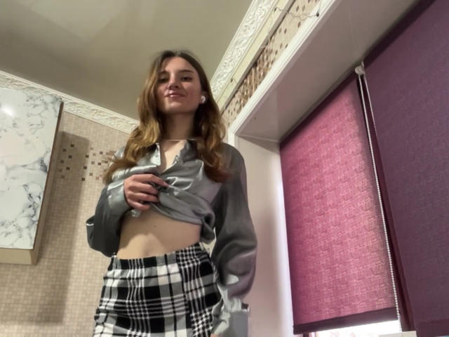 AlisaSellinger with long brown hair, lifting gray shirt to reveal flat stomach, wearing black-and-white plaid skirt, smiling slightly, standing in room with purple window shade.