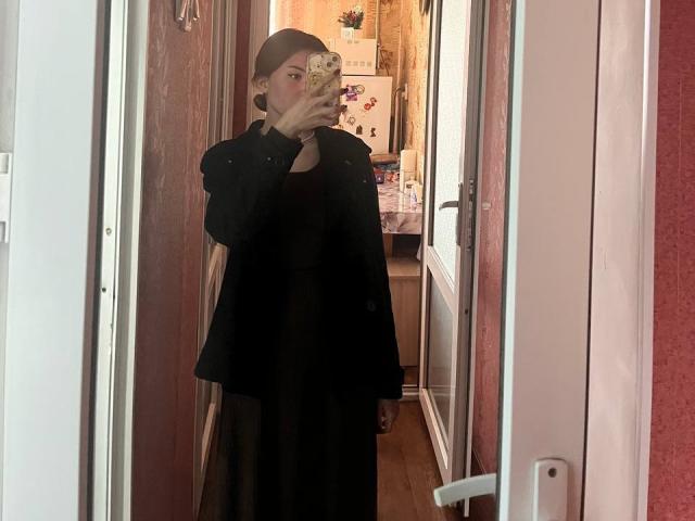 AlisaSellinger taking a mirror selfie, wearing a black coat and dress, holding a phone with a floral case, red walls and white door in the background.