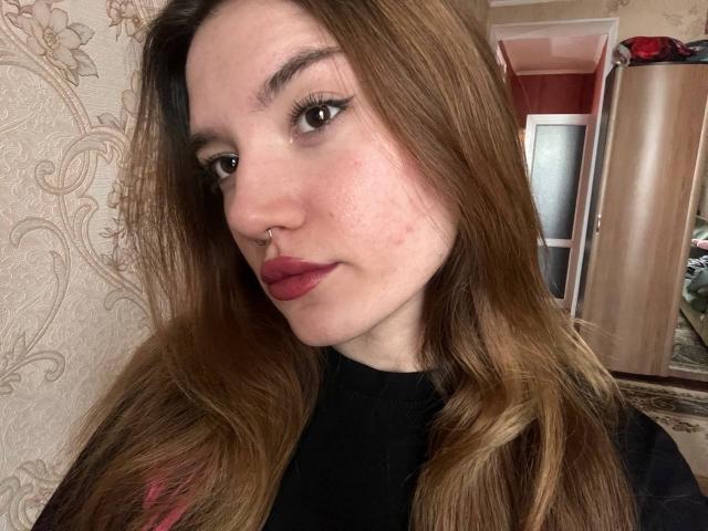 AlisaSellinger has long brown hair, red lipstick, a nose ring, and wears a black shirt. Her face shows some acne. She's looking at the camera with a slight smile.
