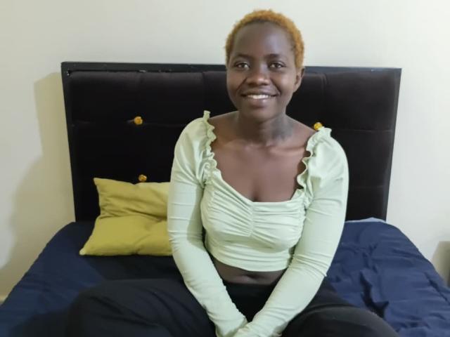 HereraPerla with dark skin, small tits, and orange hair, sitting on a bed, smiling, wearing a green ruffled crop top and black pants.