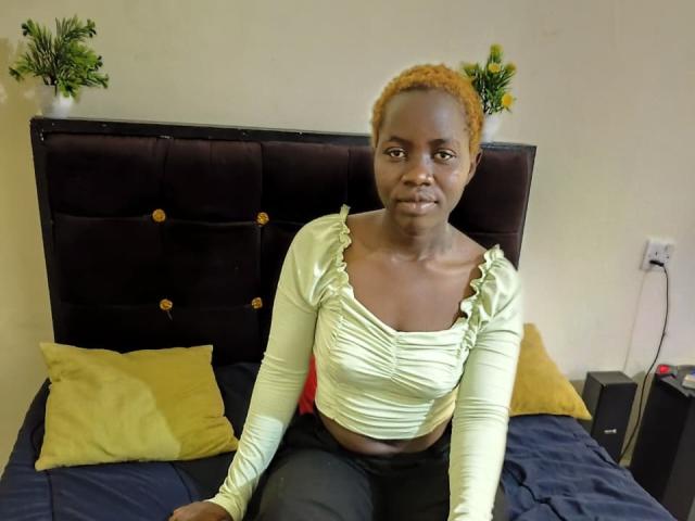 HereraPerla has dark skin, short yellow hair, small tits, and wears a light green ruffled top, sitting on a bed with yellow and green pillows.