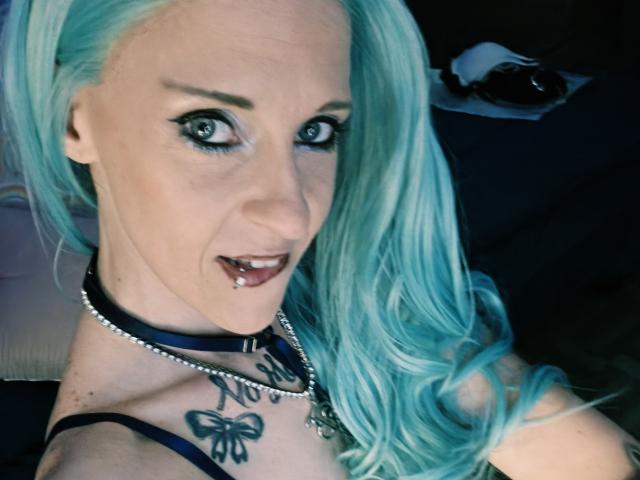 PebsStayThnx with blue hair, tongue piercing, and black collar, showing off butterfly tattoo on chest, with wide-eyed, sexy expression, and a silver necklace.