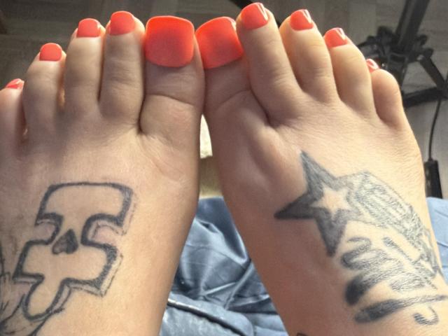 PebsStayThnx's feet with bright orange nail polish, showing tattoos: a skull on the left ankle and a star with text on the right ankle.